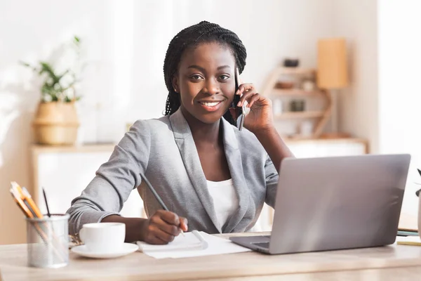 Fekete női vállalkozó beszél mobiltelefonon, jegyzetel és mosolyog a kamerába — Stock Fotó
