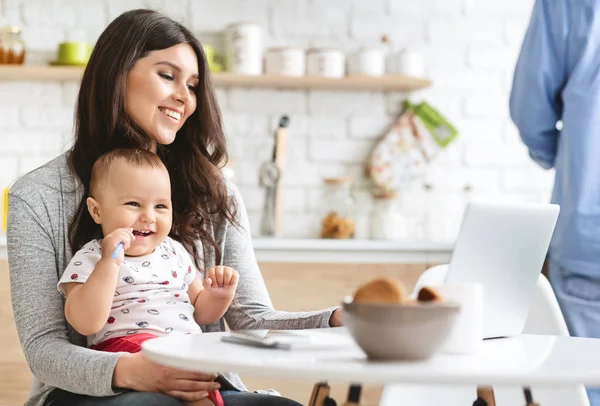 SuMillennial mother working on laptop with baby at kitchen — Stock Photo, Image