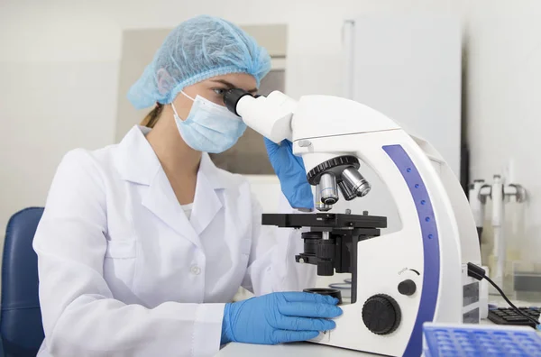 Jovem assistente de laboratório em máscara olhando através do microscópio moderno — Fotografia de Stock