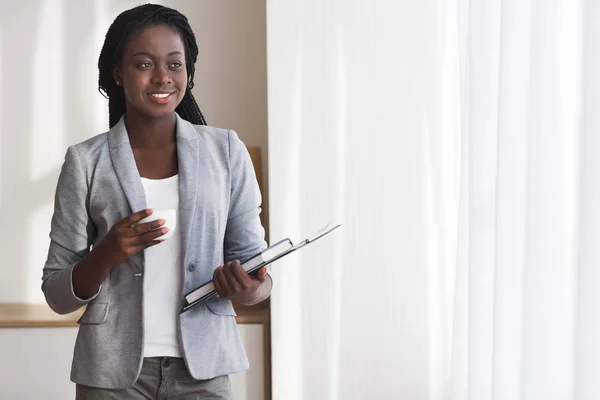 Pensive Afrikaanse secretaresse met koffiepauze in de buurt van raam in kantoor — Stockfoto