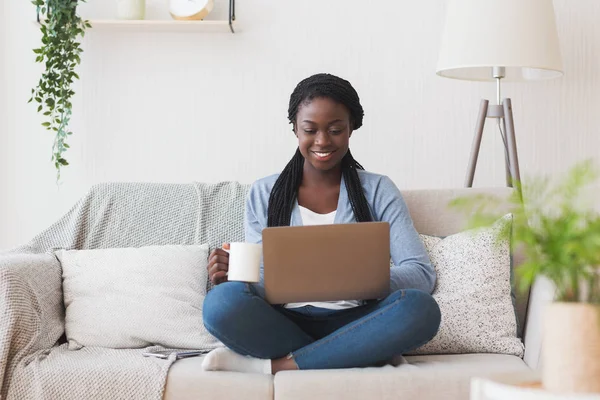 Νεαρή Αφρο κορίτσι που εργάζεται με φορητό υπολογιστή στον καναπέ στο σπίτι — Φωτογραφία Αρχείου