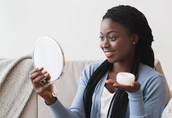 Menina afro bonita olhando no espelho de aumento e segurando creme Jar — Fotografia de Stock