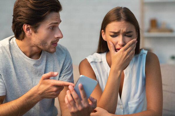 Jealous Boyfriend Showing Phone To Girlfriend Sitting On Couch Indoor