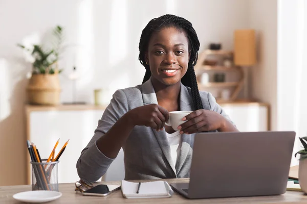 Secretária afro-americana sorrindo bebendo café no local de trabalho no escritório moderno — Fotografia de Stock