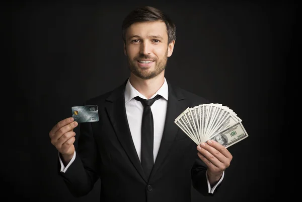 Empresario sosteniendo tarjeta de crédito y dinero de pie sobre fondo negro — Foto de Stock