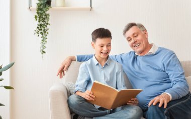 Büyükbaba, torunuyla kitap okuyor. Evdeki koltukta oturuyor.