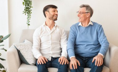 Yaşlı baba ve yetişkin oğul evde koltukta otururken gülümsüyor.