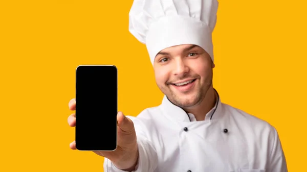 Chef mostrando tela em branco do telefone em pé no fundo do estúdio amarelo — Fotografia de Stock