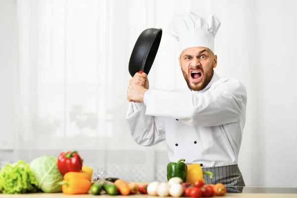 Naštvaný kuchař muž zvedá Pan hrozivě stojící v kuchyni — Stock fotografie