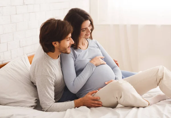Feliz esperando casal sentado na cama em casa, desfrutando de futura paternidade — Fotografia de Stock