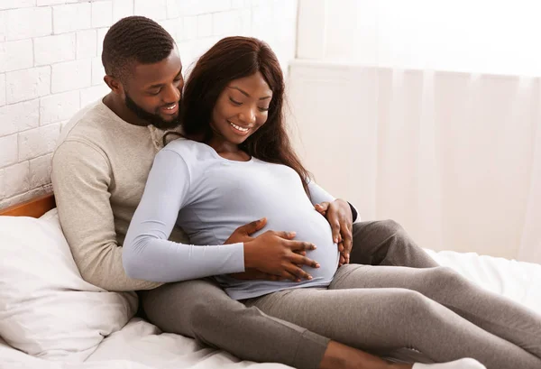Feliz jovem grávida afro casal abraçando na cama em casa — Fotografia de Stock