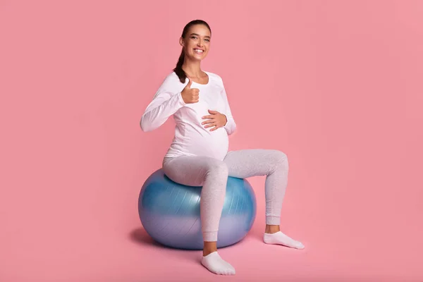 Ragazza incinta seduta su Fitball gesticolando Pollici su sfondo rosa — Foto Stock