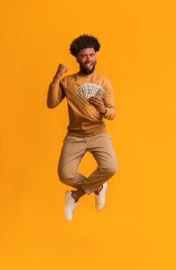 Turuncu arka planda parasıyla atlayan mutlu genç afro adam.