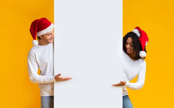 Cheerful Interracial Couple In Santa Hats Pointing At White Billboard — Stock Photo, Image