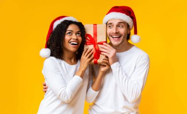 Portrait of happy couple holding gift box and wearing santa hats clipart