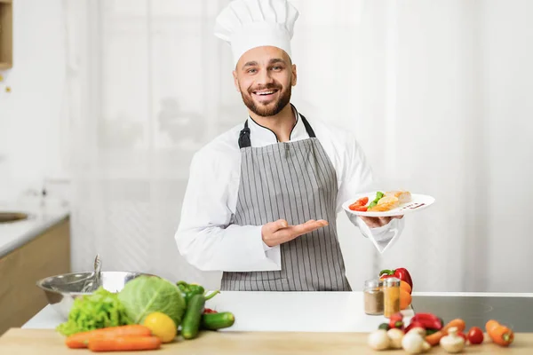 Chef uomo che tiene piatto presentando piatto in piedi in cucina coperta — Foto Stock