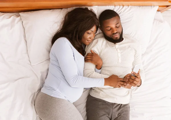 Pregnant couple resting in bed, watching photos on smartphone — Stock Photo, Image