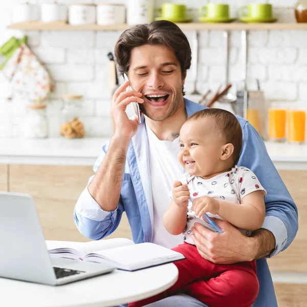 Cheerful father working at home with baby son, talking on phone