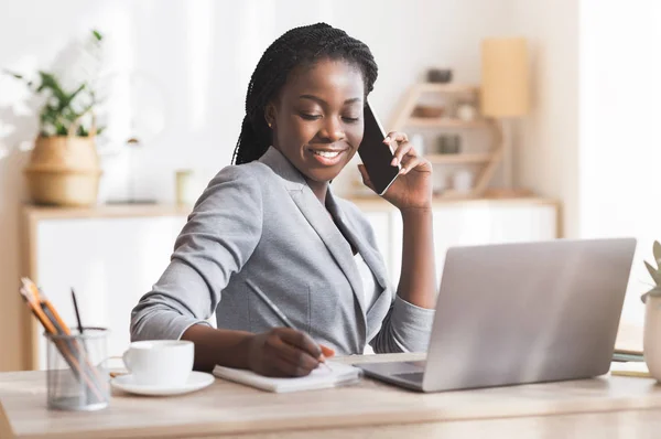 Afro imprenditrice prendere appunti pur avendo conversazione telefonica in ufficio moderno — Foto Stock