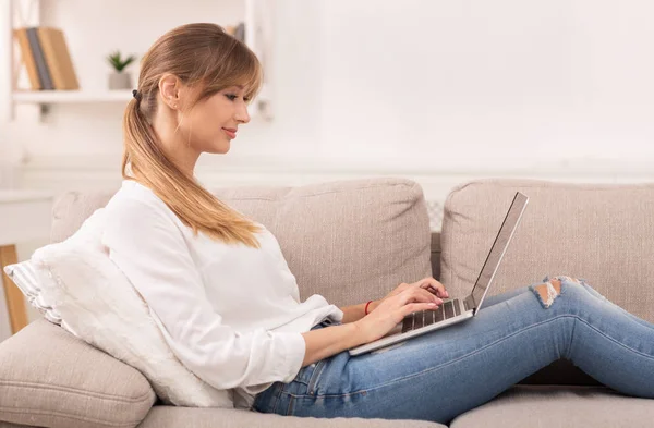 Frau arbeitet am Laptop und schreibt Artikel auf Sofa im Innenraum — Stockfoto