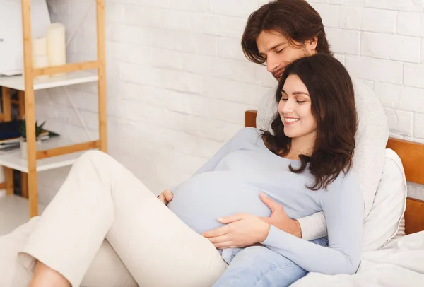 Счастливая беременная пара обнимается и отдыхает дома на кровати — стоковое фото
