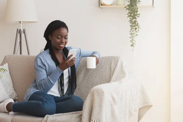 Wesoły czarny kobieta za pomocą smartfona i picia kawy na kanapie — Zdjęcie stockowe