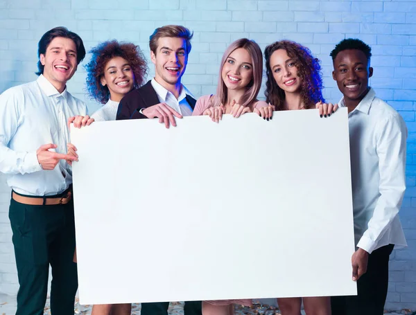 Мультиэтнические молодые люди держат белый плакат, стоящий в помещении, макет — стоковое фото