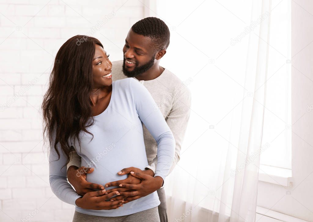 Pregnant afro couple embracing next to window at home