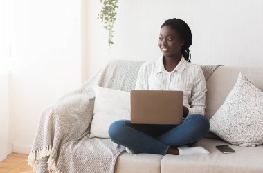 Kanepede dizüstü bilgisayarıyla oturan Afro kadın, evden internet üzerinden çalışıyor.