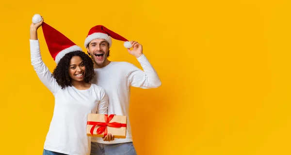 Glad interracial par som håller julklapp och leka med sina Santa hattar — Stockfoto