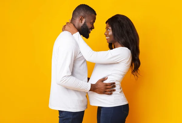 Беременная чёрная женщина и её муж обнимаются на жёлтом фоне. — стоковое фото