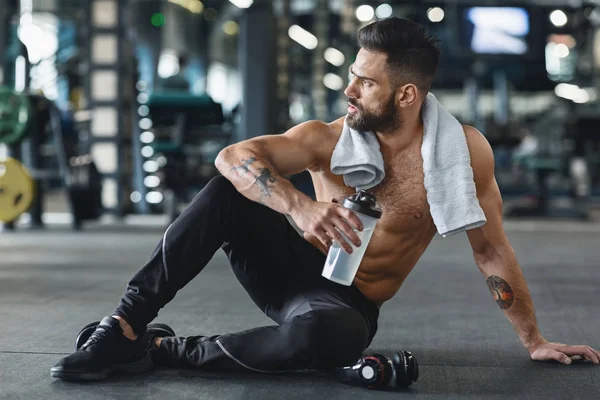 Pensive bodybuilder με νερό κάθεται στο πάτωμα στο γυμναστήριο — Φωτογραφία Αρχείου