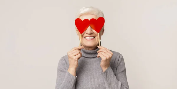 Happy aged woman covering her eyes with red paper hearts — Stock Photo, Image