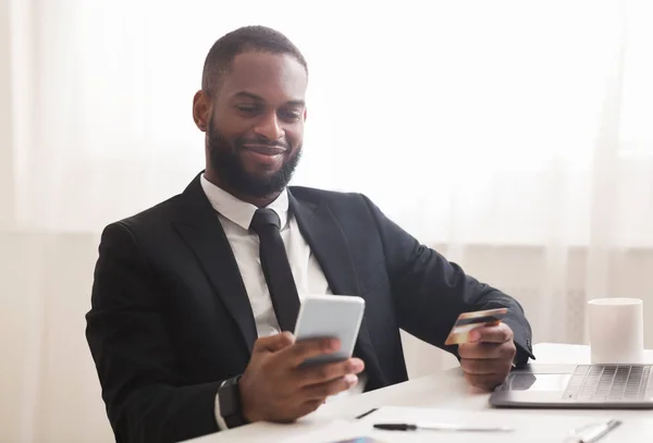 Афро - бізнесмен тримає кредитну картку, роблячи покупки в Інтернеті по телефону. — стокове фото