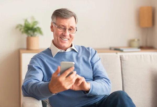 Homem idoso alegre usando celular sentado no sofá em casa — Fotografia de Stock