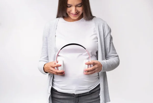 Niña embarazada sosteniendo auriculares cerca del vientre de pie, estudio, recortado —  Fotos de Stock