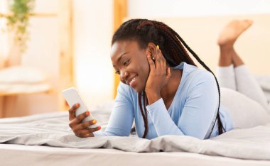 Akıllı telefon kullanan neşeli afro kadın yatakta uzanıyor.