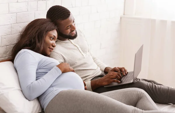 Pregnant lady and her husband using laptop, laying on bed — 스톡 사진