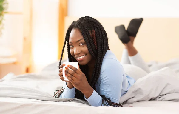 Afro chica teniendo café acostado en cómoda cama en el dormitorio —  Fotos de Stock