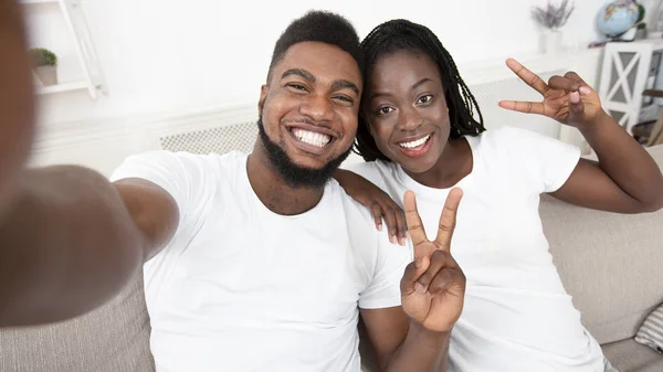 Joyful black couple taking selfie and showing peace gesture at home — ストック写真