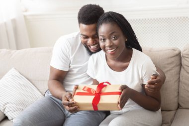 Romantik çift evde kanepede otururken birbirlerine sarılıp hediye kutusunu tutuyorlar.