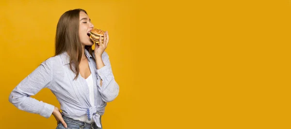 Millennial girl eating burger standing on yellow studio background, panorama — Stock Photo, Image