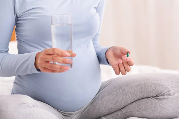 Неузнаваемая беременная женщина с таблетками и водой, сидящая на кровати — стоковое фото