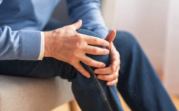 Старший менеджер масажує коліно, сидячи на дивані в приміщенні, обрізаний — стокове фото