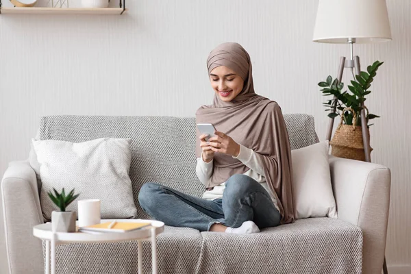 Happy arab wanita dalam jilbab menggunakan smartphone di sofa di rumah — Stok Foto
