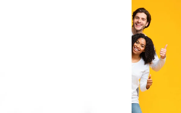 Couple multiracial regardant derrière le panneau publicitaire blanc et montrant pouces vers le haut — Photo