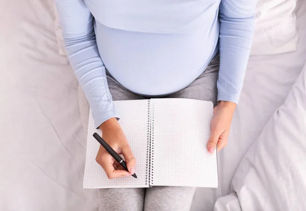 Unerkennbar schwangere Frau macht sich Notizen im Bett sitzend drinnen, Draufsicht — Stockfoto