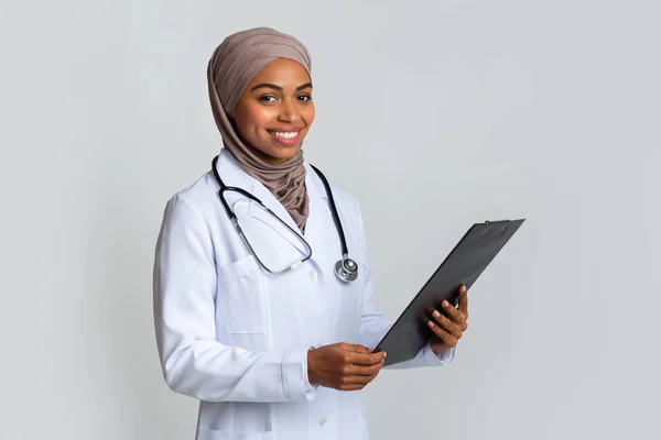 Smiling black muslim female doctor in hijab with clipboard in hands