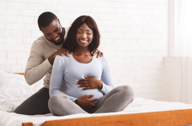 Afro adam hamile karısına masaj yapıyor, yatakta oturuyor.