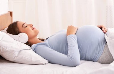 Yatakta uzanıp müzik dinleyen sakin hamile kız.